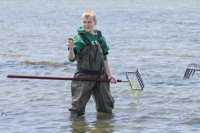 a teenage boy wearing waders holding a quahog and a clam rake