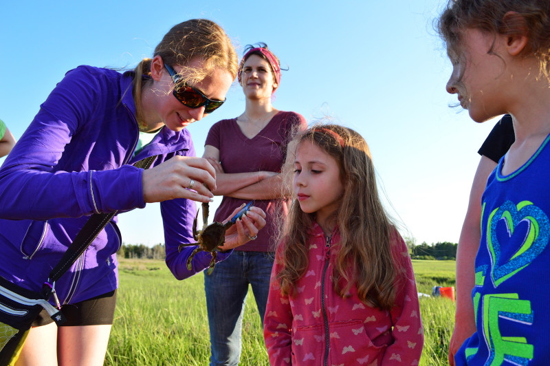 a woman holds up a blue crab to show two young girls