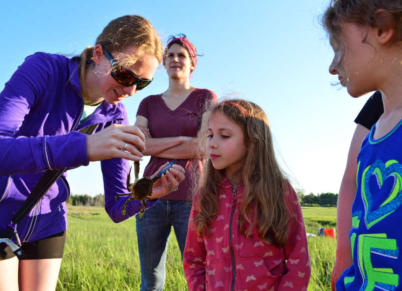 a woman holds up a blue crab to show two young girls