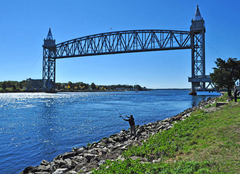 a man fishes from the shore of Cape Cod Canal next to the railroad bridge in Bourne