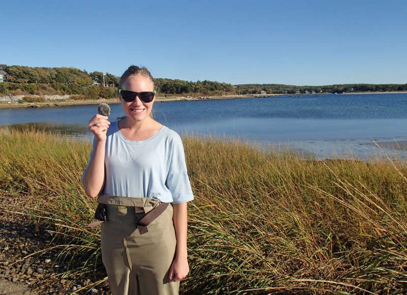 a woman holds up a perfect bay scallop shell next to West Falmouth Harbor