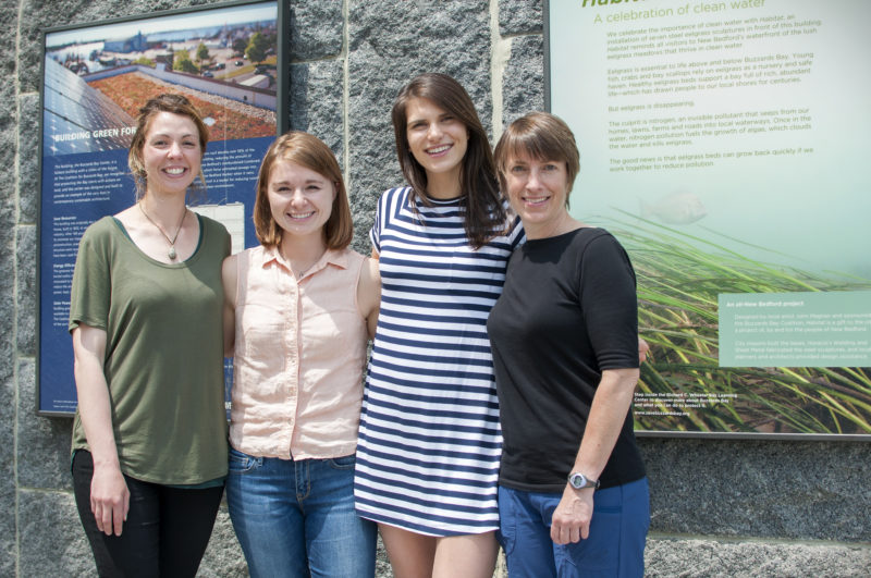 Four new staff members standing outside the Coalition’s stone building, with signs about green energy and clean water in the background.