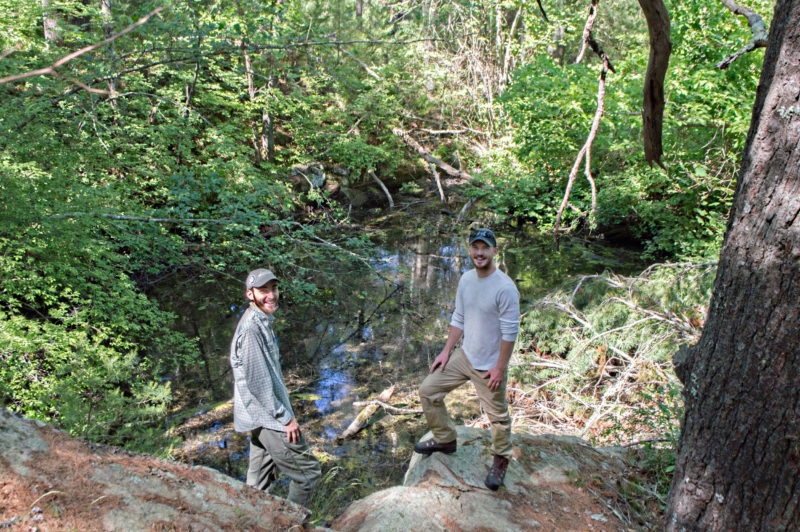 Two young men beside a vernal pool surrounded by green trees