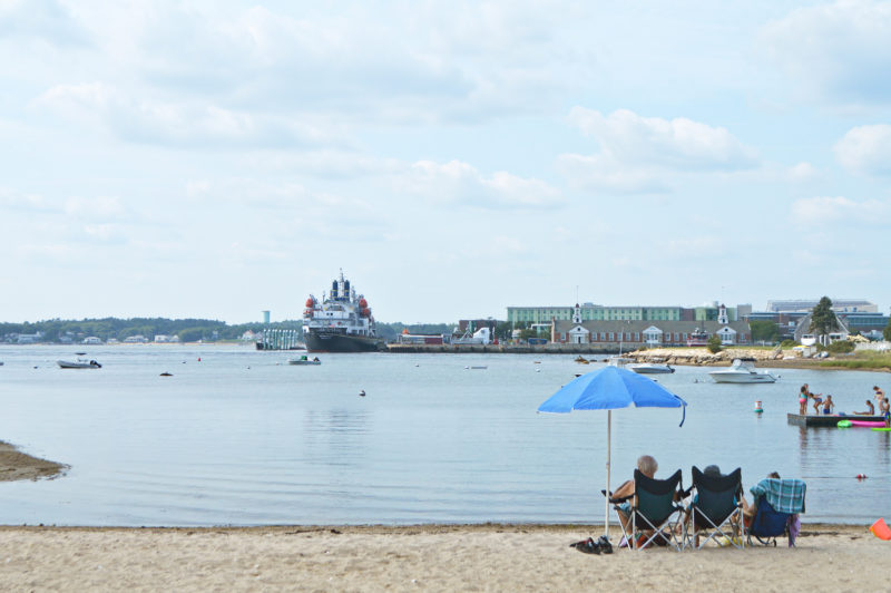 People beneath a beach umbrella look out at a boat in the Cape Cod Canal from Gray Gables Beach in Bourne