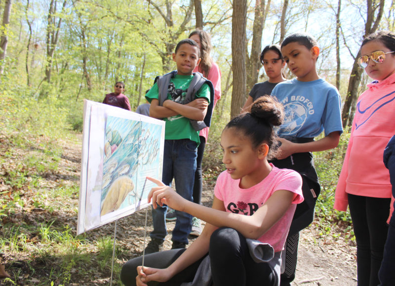 A group of kids stands in front of a story page as one girl points out a line