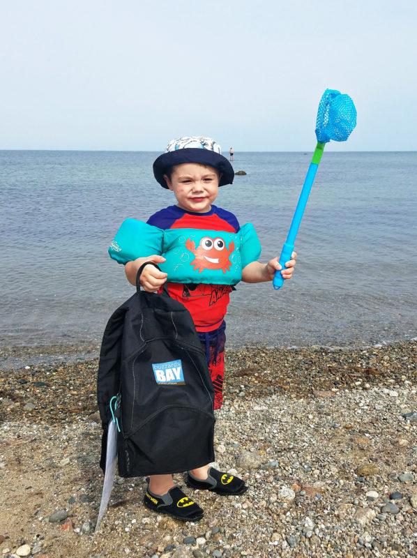 A young boy holding a BBC backpack and a fishing net on a beach