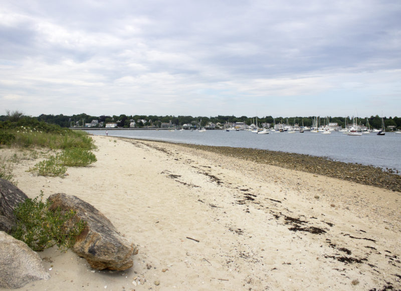 The sandy shore of Knowles Beach in Dartmouth, with the sailboats of Padanaram Harbor in the background